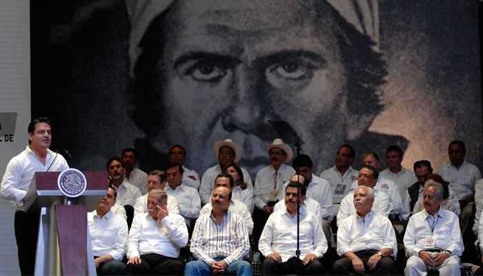 Foto del gobernador Aristóteles Sandoval dando un discurso, con varias personas sentadas a su lado y la imagen del rostro de Morelos al fondo