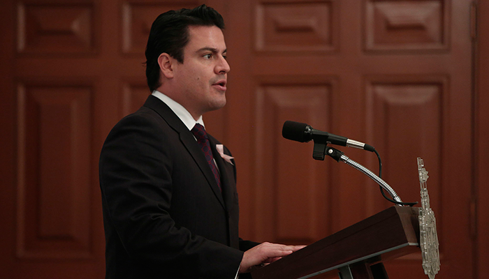 Foto de perfil del gobernador hablando en un podium con micrófono, al fondo una pared de madera tallada con rectangulos alargados